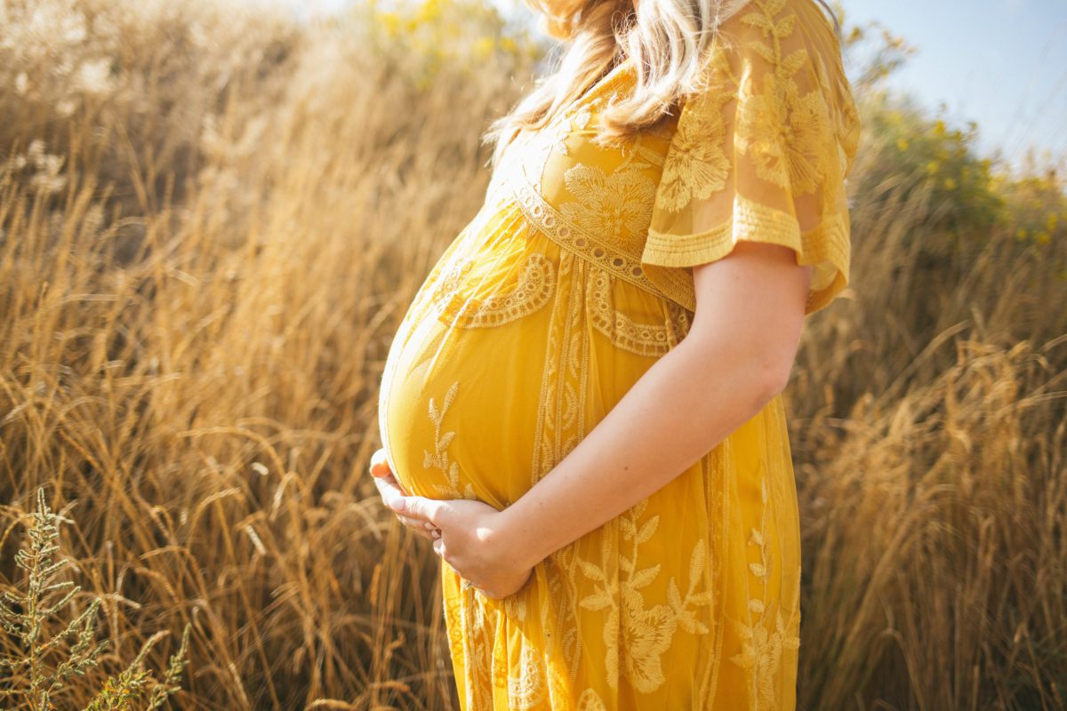 Pregnant woman wearing a yellow dress in front of field.