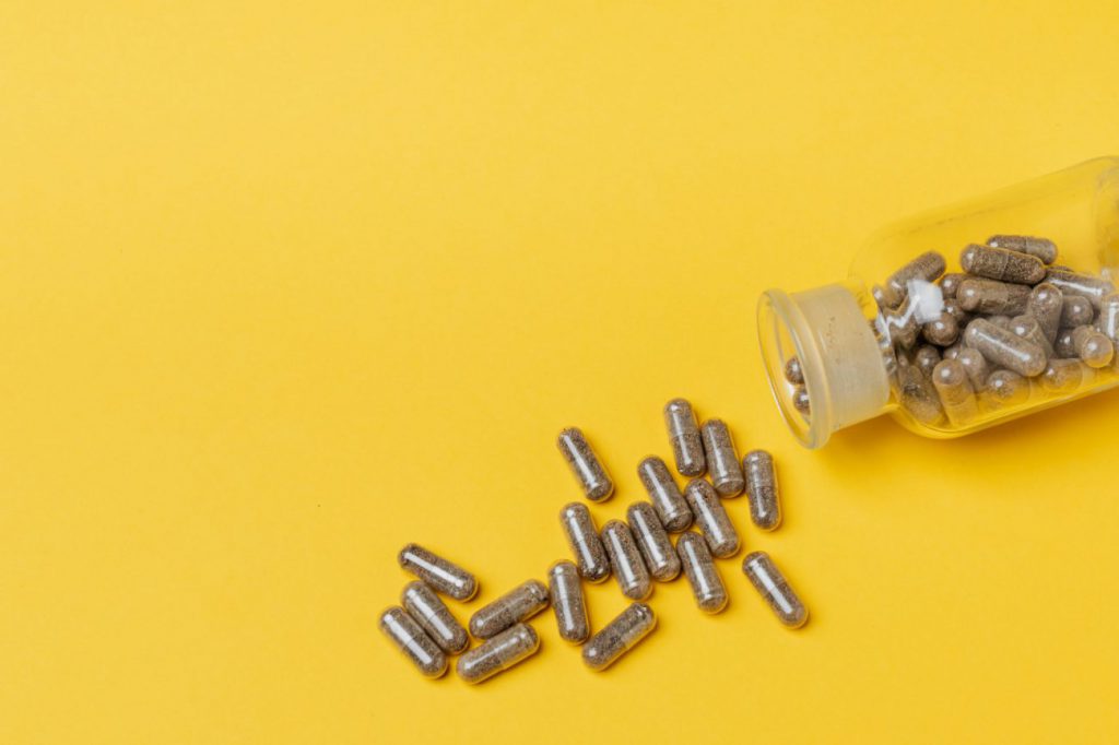 Spilled bottle of grey supplement capsules on yellow backdrop.