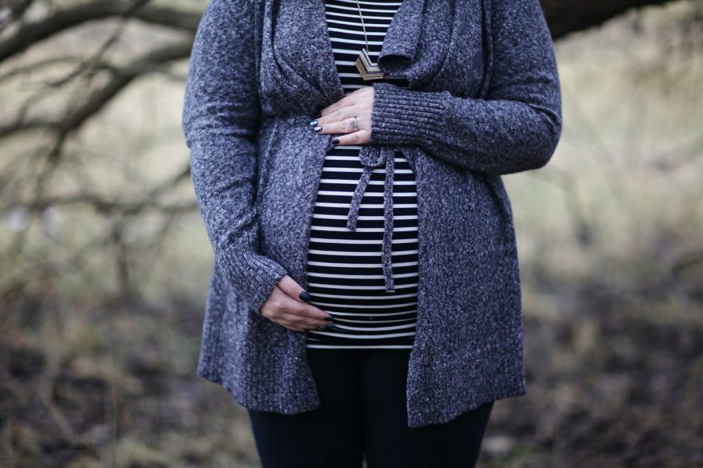 Pregnant woman standing outside wearing a grey sweater and black and white striped shirt holding her belly.